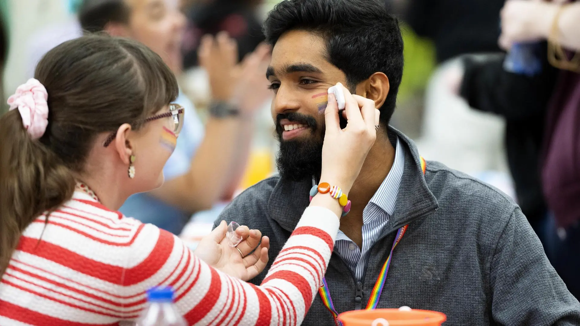 A person is getting a pride flag painted on their cheek by a student at a Pride fair.