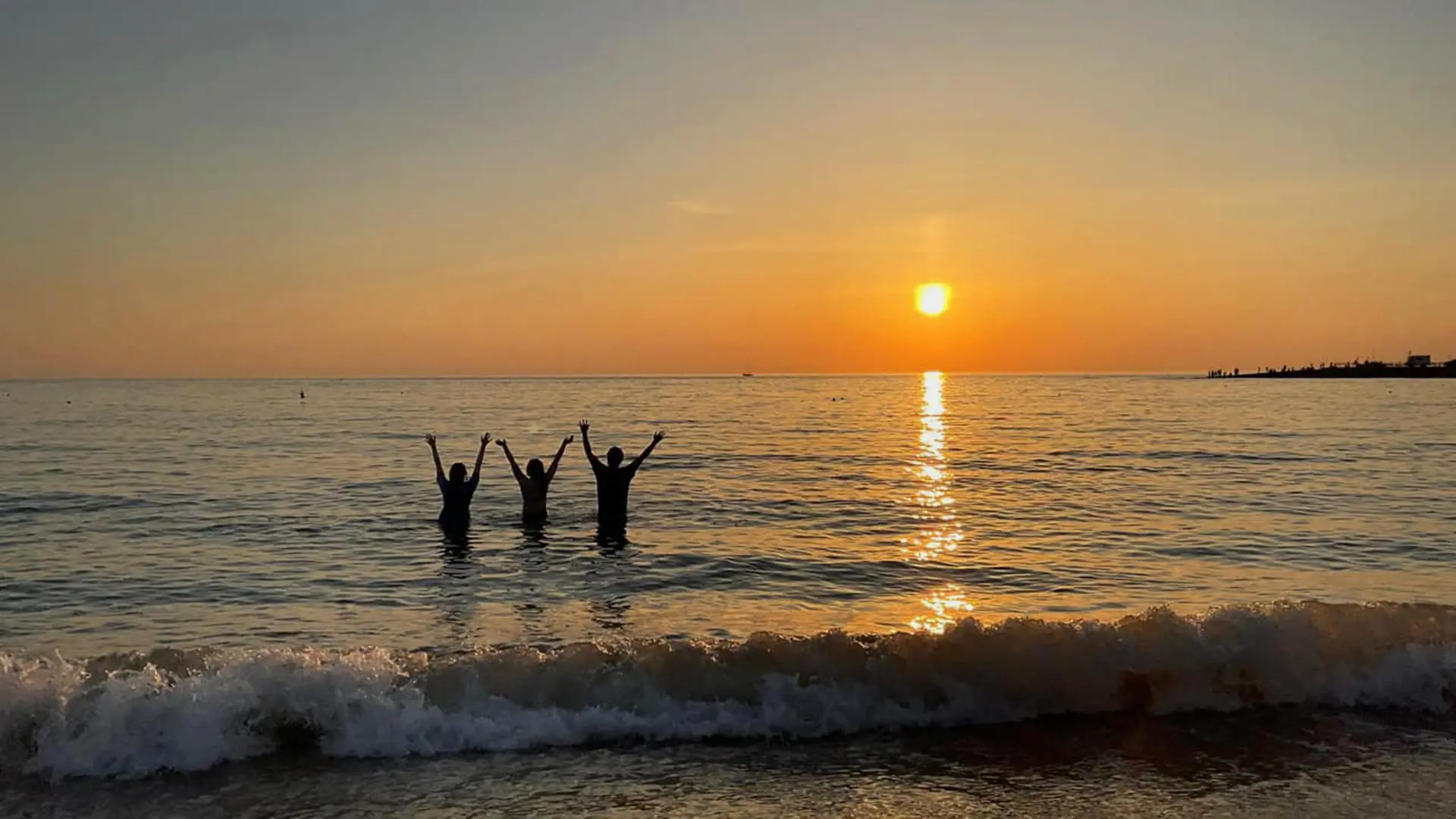 A group stand in the sea at a beach as the sun sets on the horizon