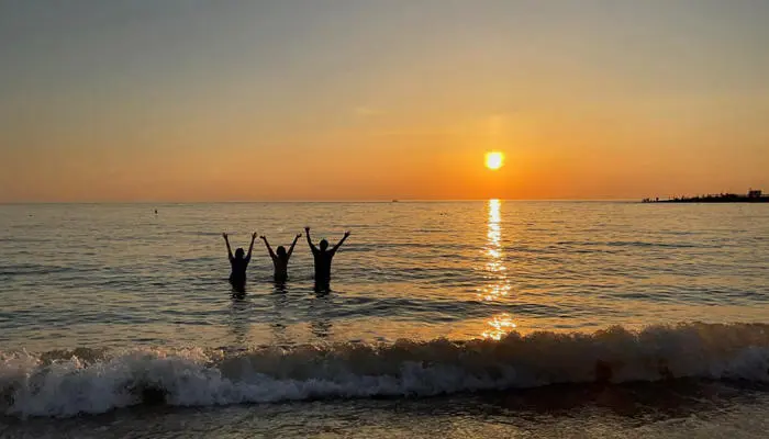 A group stand in the sea at a beach as the sun sets on the horizon