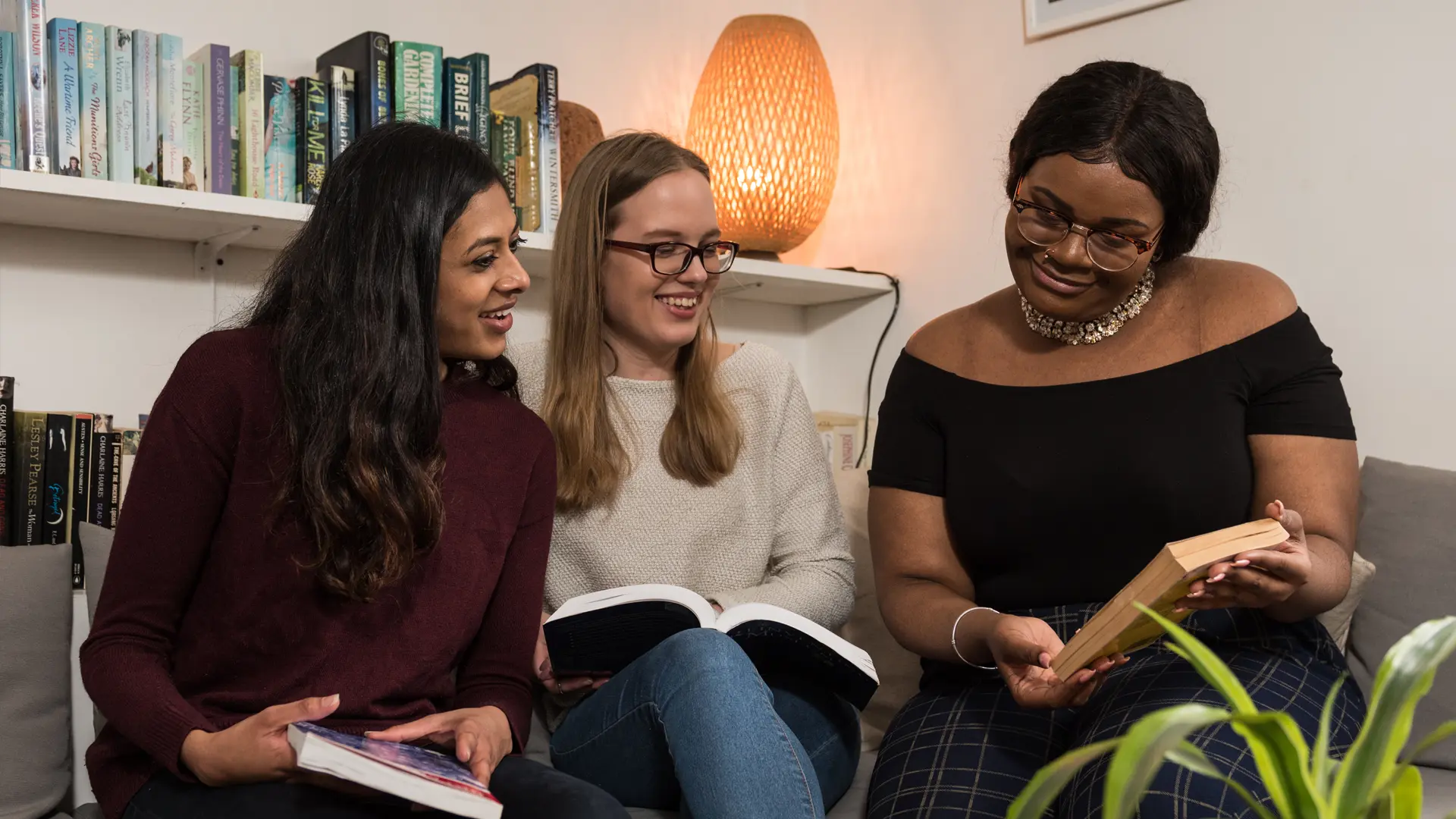 Three female students sat in a living room on a sofa looking at a book