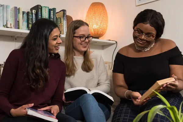 Three female students sat in a living room on a sofa looking at a book
