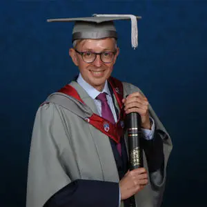 Mick Cartledge dressed in graduation gown holding an Honorary Fellow award