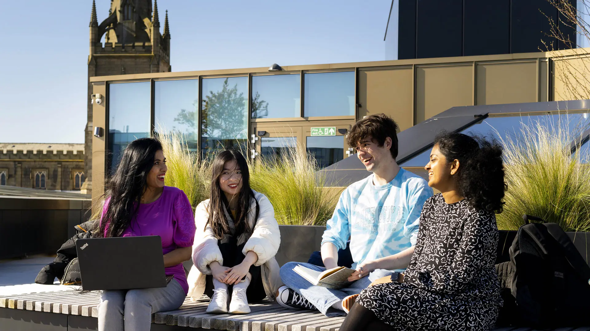four international students sat on the roof terrace of the student centre on a bench on a sunny day