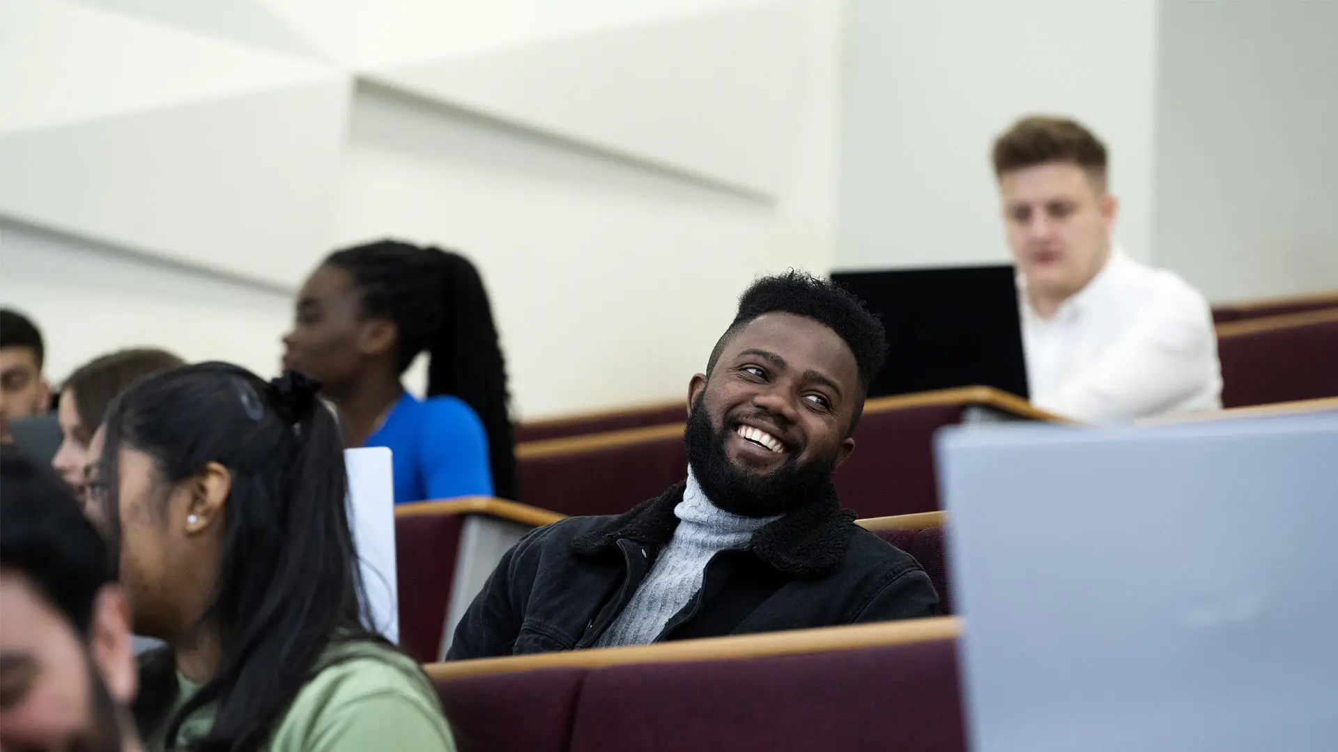 group of students sat in a lecture theatre using laptops, main focus is a student wearing a black denim jacket and grey turtle neck - smiling and looking away from the camera