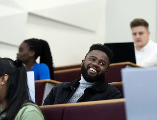 group of students sat in a lecture theatre using laptops, main focus is a student wearing a black denim jacket and grey turtle neck - smiling and looking away from the camera