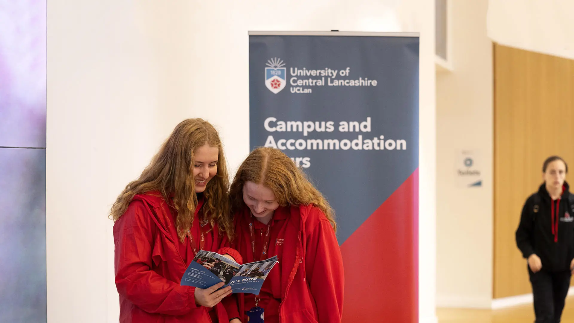 two student ambassadors in red stood in front of a sign advertising campus and accommodation tours