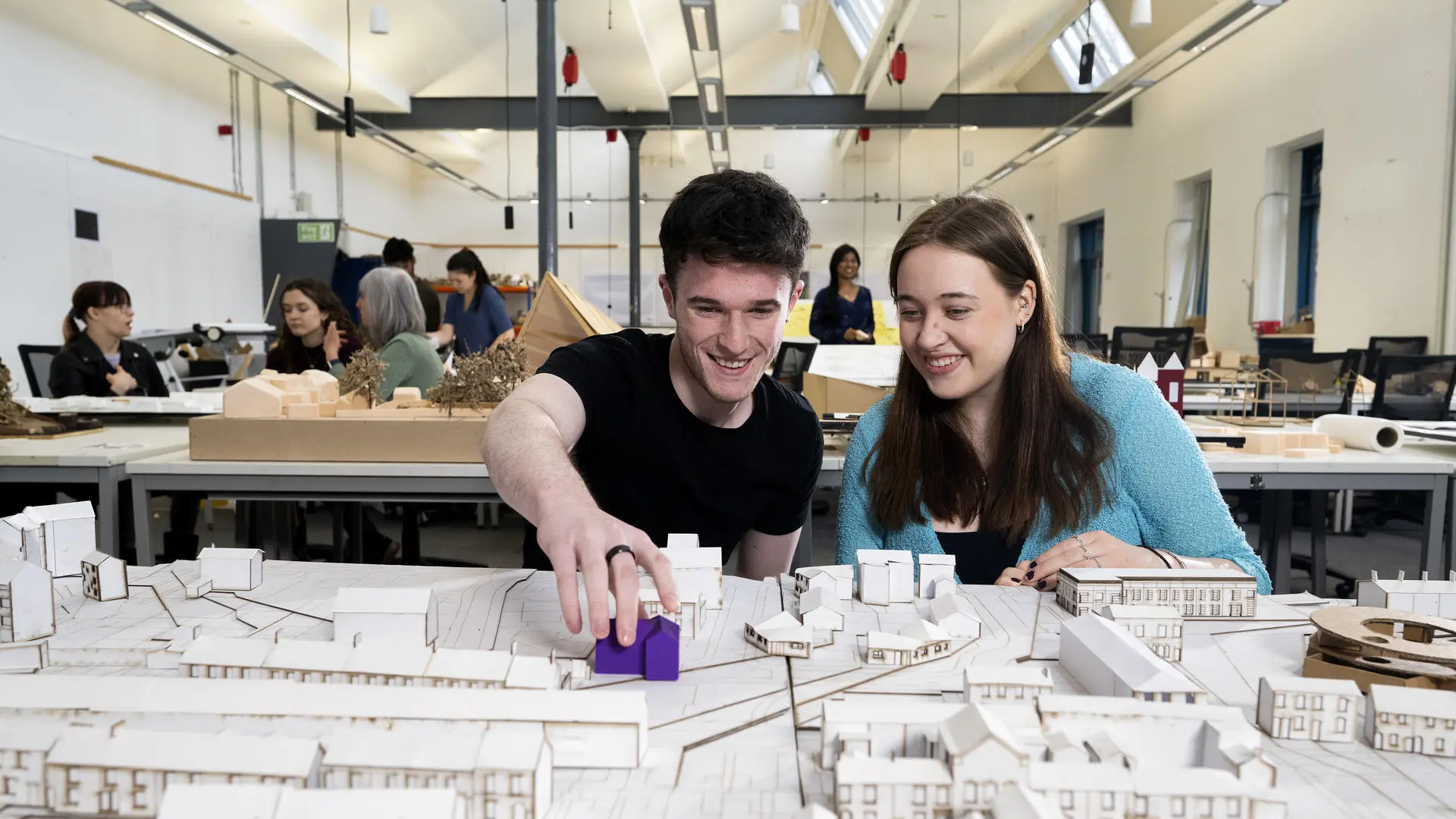 two students looking at model houses in an architecture studio with other students working in the background