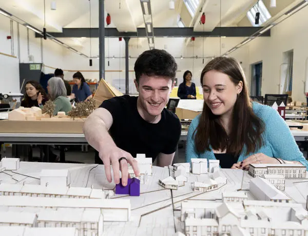 two students looking at model houses in an architecture studio with other students working in the background