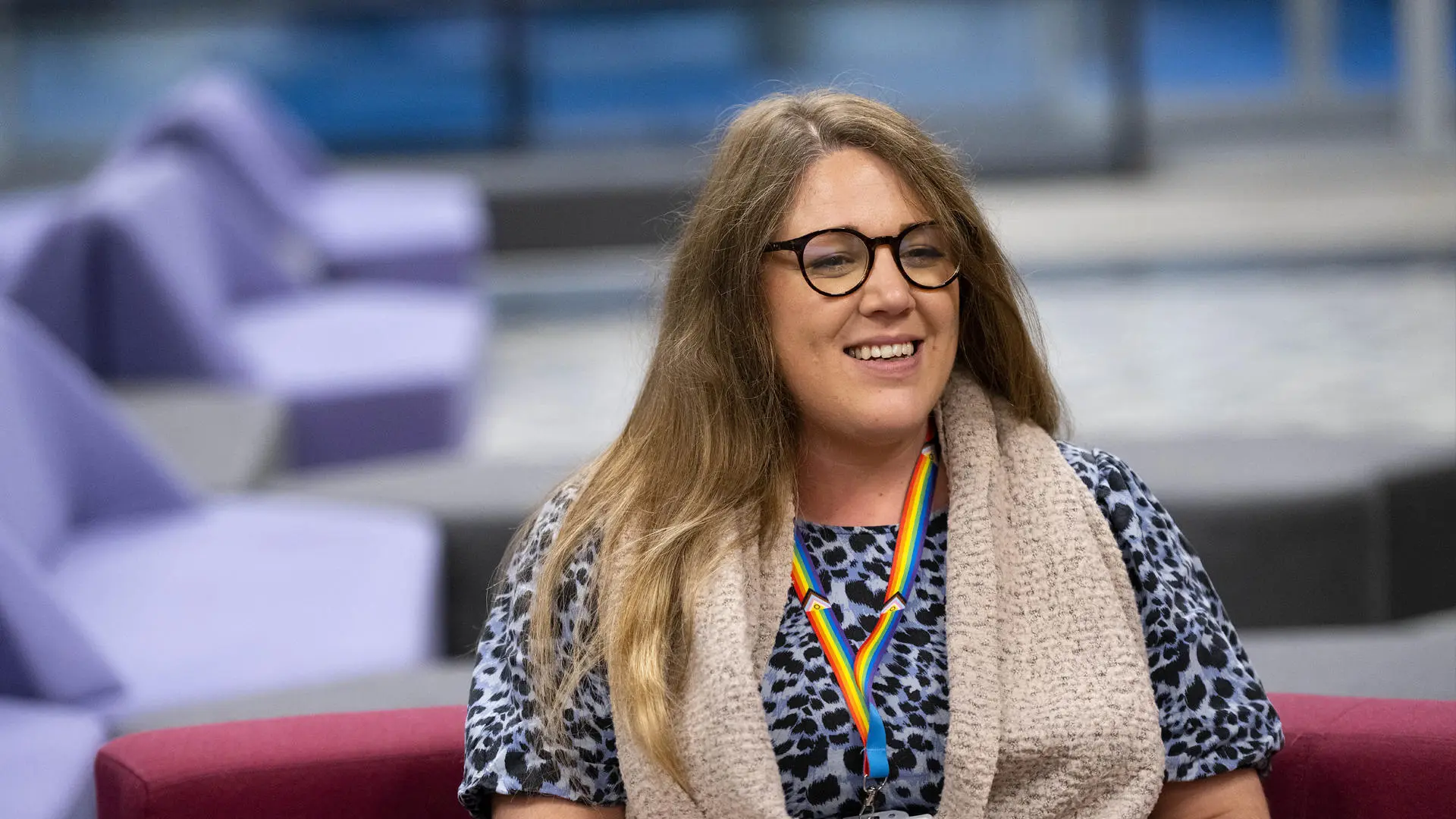 harriet tyler from the people team sat on a burgundy sofa with a blue leopard print top on, glasses and a rainbow lanyard
