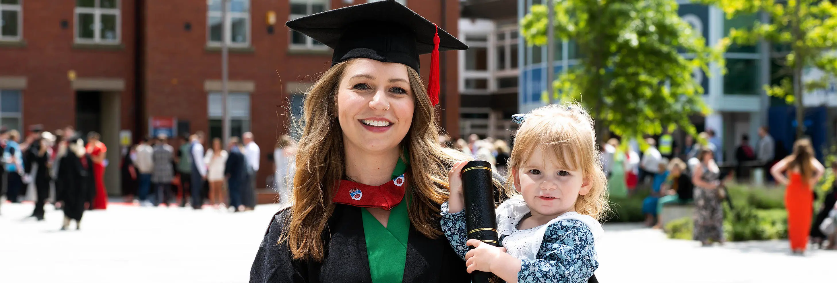 Image of UCLan graduate Emily Chell, in black graduation cap and gown, holding young child with background of buildings and trees