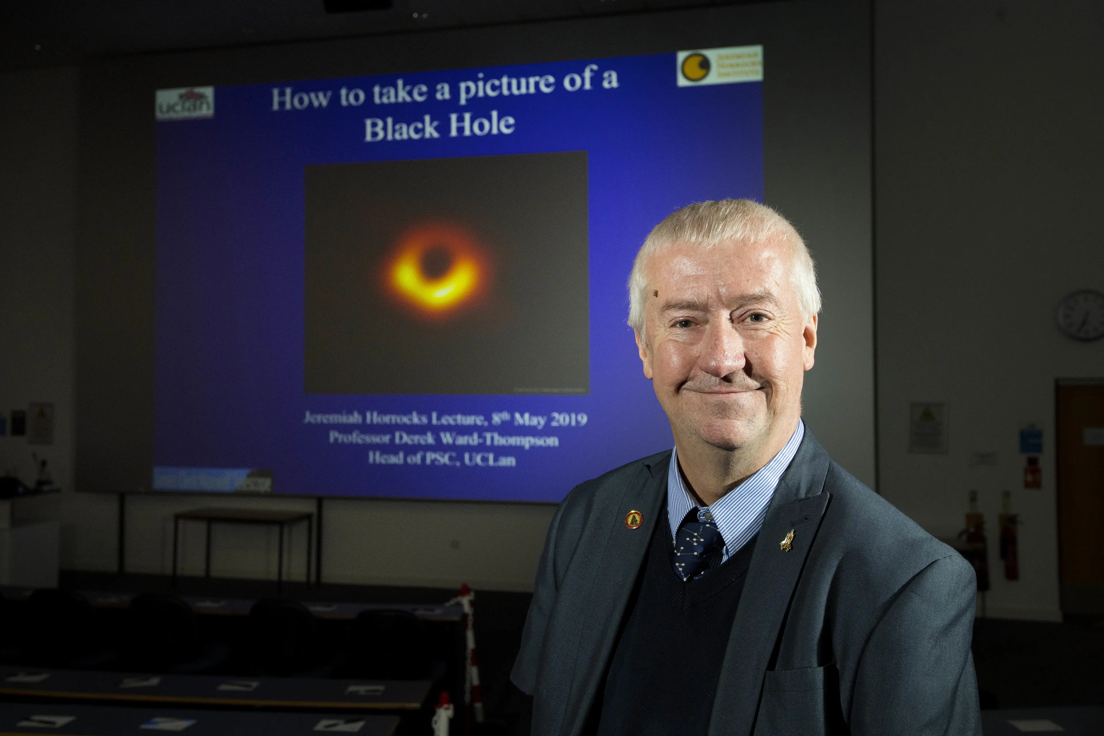 Professor Derek Ward-Thompson sat in front of a large lecture theatre screen which shows a photo of a black hole